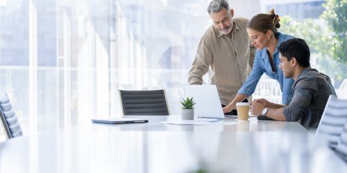 Group of business people working. The team are standing and sitting at a board room table looking at a laptop computer. All dressed in casual business clothing with a window behind them